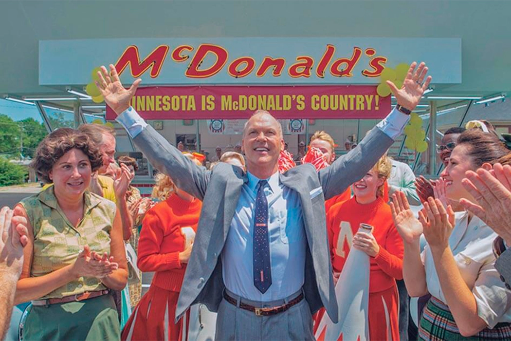 Michael Keaton no filme fome de poder. Ator está de terno e gravata, com pessoas felizes ao fundo da empresa Mc Donalds