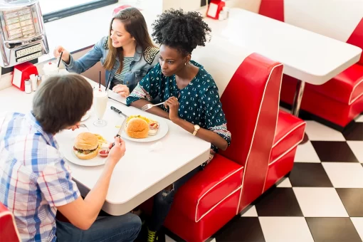 Amigos em lanchonete, duas mulheres e um homem apreciando hamburguer. O local tem cadeiras vermelhas, mesas brancas e chão quadriculado em preto e branco, dando uma ideia de um local mais depochado, frequentados por jovens e adultos.